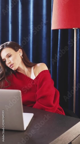 Sensual Young Woman in Red Sweater and Earbuds Working on Laptop, Looking at Screen with Soft Dreamy Smile, Tilting Head While Sitting at Desk in Cozy Room with Blue Curtains and Lamp