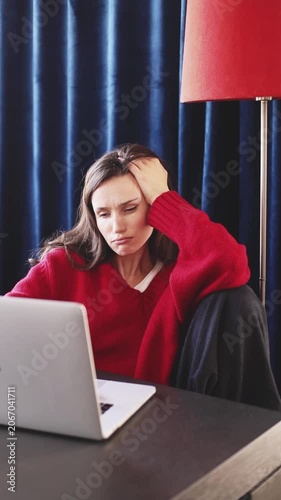 Bored and Disappointed Woman Looking at Laptop Screen, Holding Hand on Head and Making Grumpy Face expression, Sitting in Cozy Room with Red Floor Lamp and Dark Blue Velvet Curtains