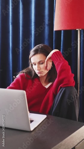 Exhausted Bored Woman in Red Sweater Sitting at Desk and Looking at Laptop Screen, Feeling Sleepy, Touching Face and Eye Rubbing from Fatigue in Room with Dark Blue Curtains and Floor Lamp