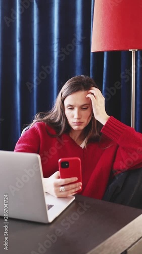 Sad Tired Woman Sighing and Pouting While Looking at Smartphone Screen, Holding Head in Hand, Feeling Overwhelmed and Disappointed at Desk near Laptop in Cozy Dark Blue Room with Red Lamp