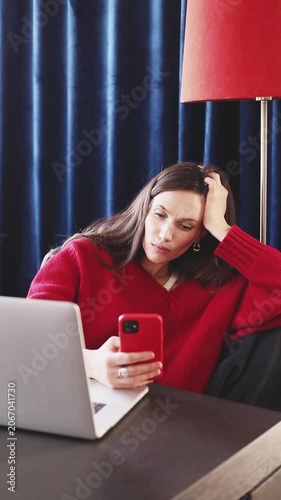 Tired Bored Woman in Red Sweater Sitting at Desk with Laptop and Looking at Smartphone, Holding Head in Hand with Exhausted Expression in Room with Dark Blue Curtains and Red Floor Lamp