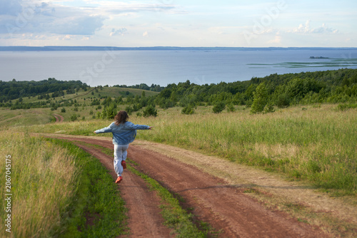 A little girl running along a picturesque winding field road leading to the riverbank through a meadow. Summer.