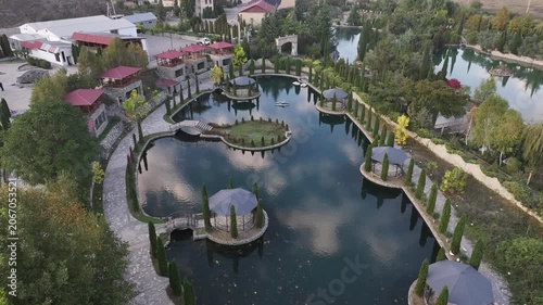 An aerial drone view showing a beautiful resort park with a calm lake, elegant gazebos, stone pathways, and lush cypress trees reflecting in the water during sunset.

