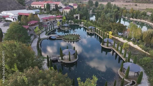 An aerial drone view showing a beautiful resort park with a calm lake, elegant gazebos, stone pathways, and lush cypress trees reflecting in the water during sunset.

