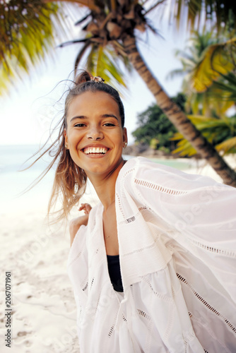 Happy woman with flowing hair smiling on tropical beach with palm trees in white summer outfit enjoying paradise vacation. Summer holidays selfie. Generative AI.