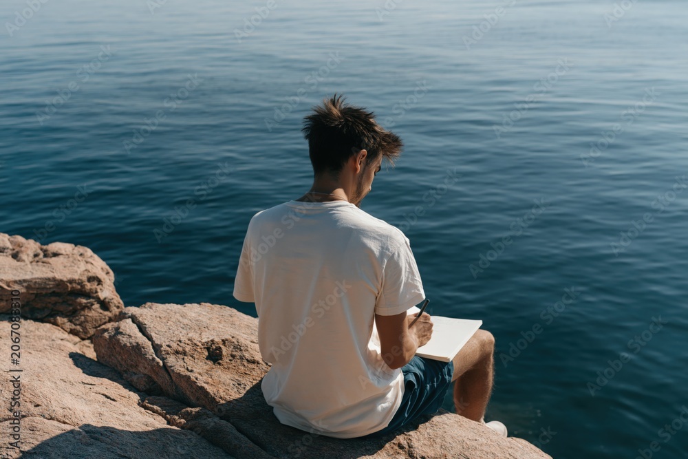 Fototapeta premium Back view of young man on rocky shore writing in notebook for ocean background and mindful travel journal or study scene