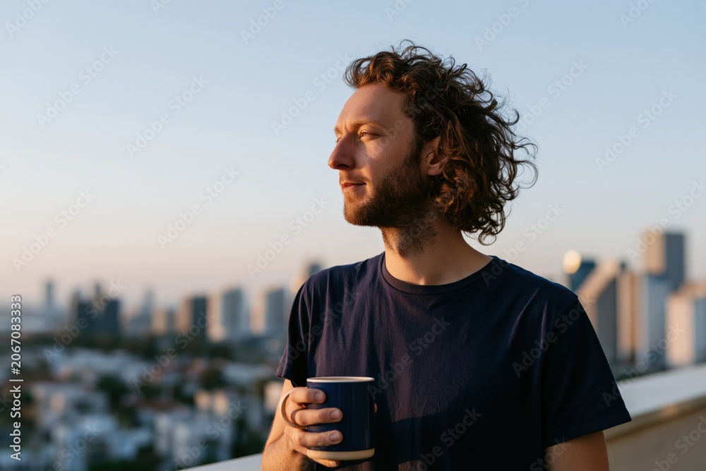 Fototapeta premium Young man holding coffee mug on rooftop at sunrise with city skyline for lifestyle banner and morning routine background