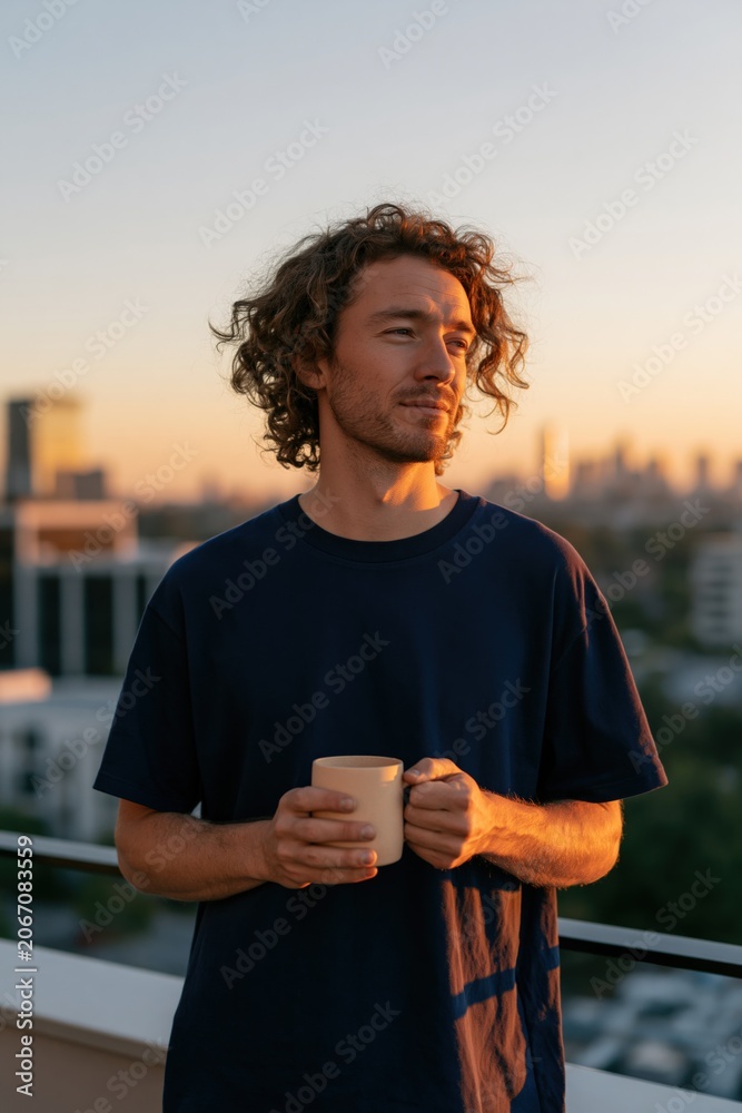 Fototapeta premium Young man with curly hair holding a warm mug on a rooftop at golden hour for lifestyle portrait and morning routine background for wellness and mindful living banner