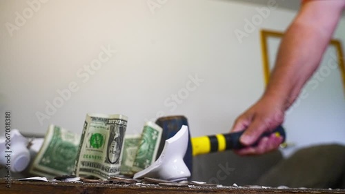 Slow motion footage showing a hand smashing a white ceramic piggy bank with a hammer. Coins and dollar bills explode from the broken pieces