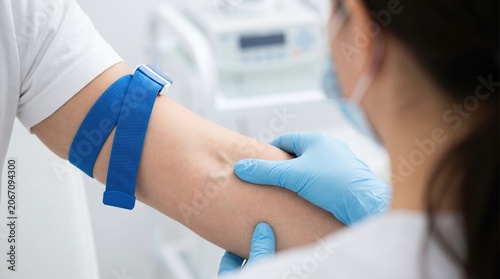 A health worker in gloves examines a patient's arm while applying a tourniquet before taking a blood sample. The clinic has medical equipment and is well lit during the day