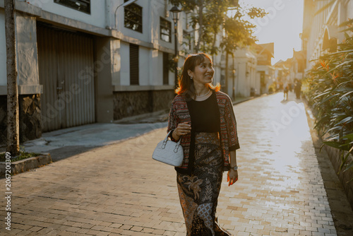 Happy Fashionable adult Asian woman in batik textile layered outfit carrying white small bag leather walking in golden hours city. Urban street style and everyday fashion culture.