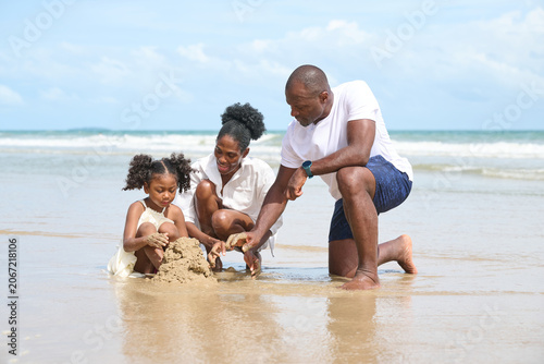 Happy family, including a father, mother, and daughter, playing together on a sandy beach next to the ocean. They are building a sandcastle under a bright, sunny sky