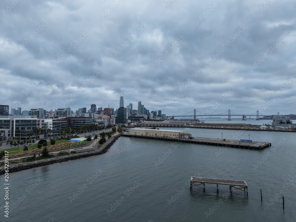 Obraz premium San Francisco skyline and Bay Bridge on a cloudy day, showing modern buildings, urban waterfront development, and industrial piers alongside Mission Bay