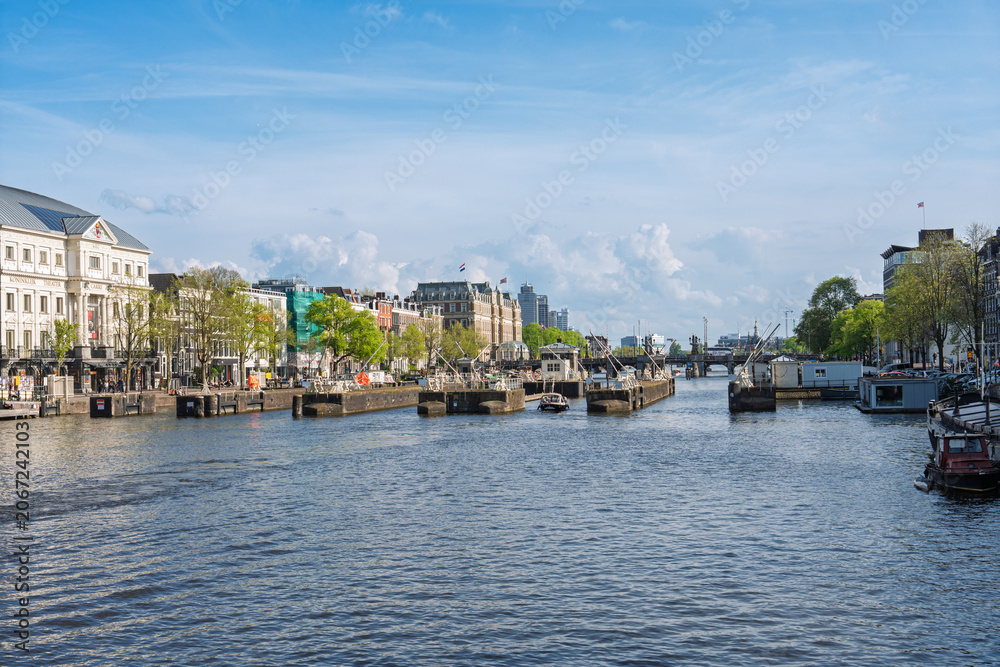 Obraz premium A beautiful sunny day view of the Amstel River in Amsterdam, Netherlands, featuring historic buildings, canal infrastructure, and calm water under a bright blue sky in spring.