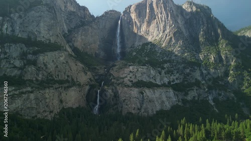 Aerial shot of Yosemite Valley featuring cascading waterfall surrounded by granite cliffs, dense forest, and dramatic mountain landscape in California’s Sierra Nevada.