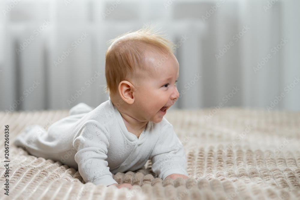 Fototapeta premium Happy baby dressed in white bodysuit lying on stomach and looking to the side, expressing joy and laughter during childhood development on a comfortable bed indoors