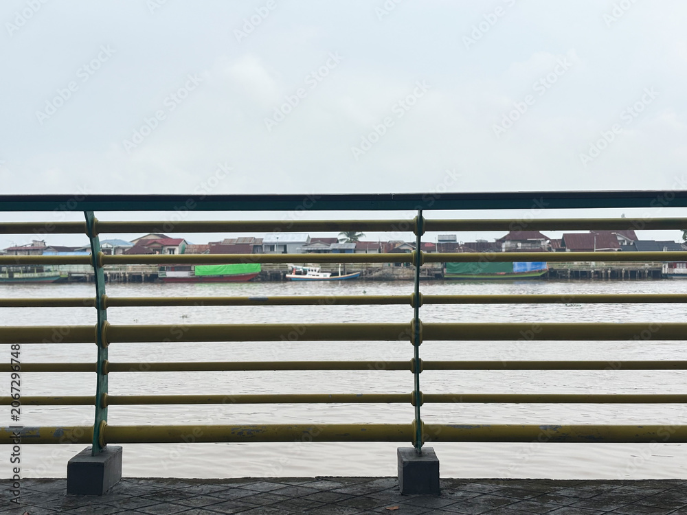 Fototapeta premium Yellow metal railing on a waterfront promenade looking over a wide river and a traditional riverside village under a cloudy sky