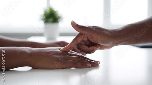 A black hand touches another hand lightly on a white desk with a blurred plant in the background, showing support and empathy