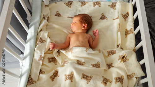 Newborn baby calmly sleeping in a white wooden crib, wrapped in a blanket with a charming teddy bear pattern, depicting innocence and early childhood