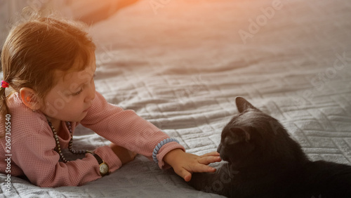 Red haired toddler gently reaches to touch black cat lying on white quilted bed under warm sunlight