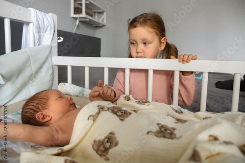 Older sister gently holding the tiny hand of her sleeping newborn baby sibling in a white crib, showing love and the beginning of their special family bond in the nursery