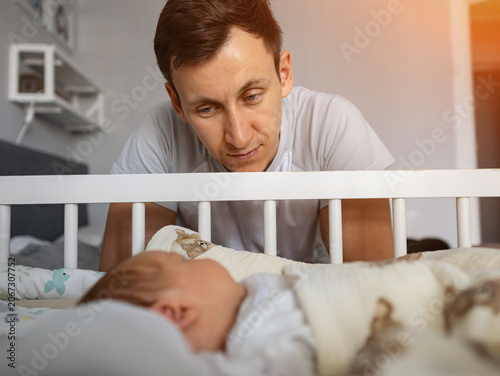 Loving father looking down at his infant child sleeping peacefully in a white crib, representing parental care, family bonding, and the tenderness of new parenthood