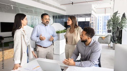 Group of professionals engaged in discussion in modern coworking office, listening opinion and ideas speaking Indian mature team leader, gathered together in contemporary corporate workspace. Briefing
