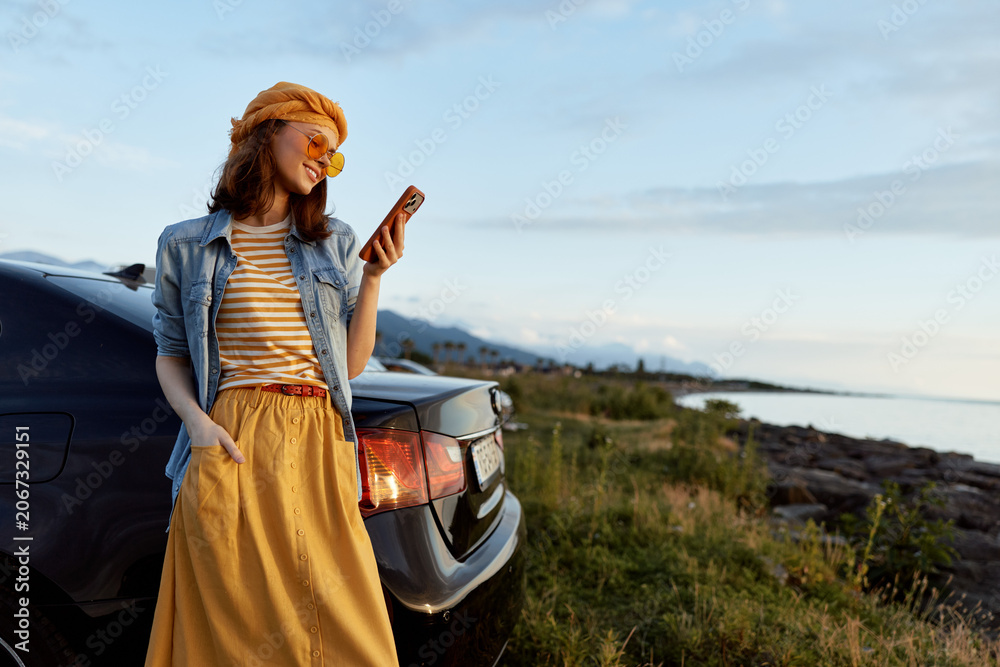 Fototapeta premium Cheerful woman outdoors, wearing striped top and yellow skirt, leaning against a car by the coast, enjoying a sunset moment, casual lifestyle, natural colors