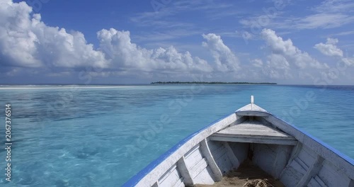 POV from the bow of a wooden boat cruising across calm turquoise tropical ocean waters, sunny summer vacation scenery with blue sky and white clouds.