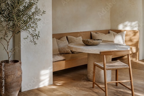 Minimalist dining nook featuring a built-in wooden bench, round stone table, woven chair, and a potted olive tree