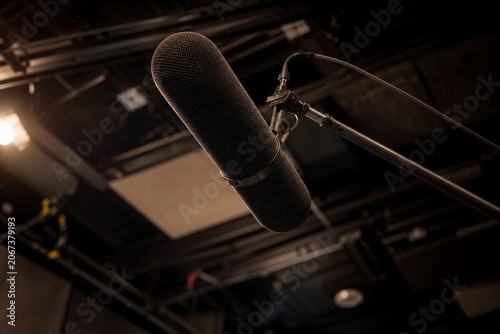 Shotgun microphone with a black blimp windscreen on a boom pole inside a dark film production soundstage