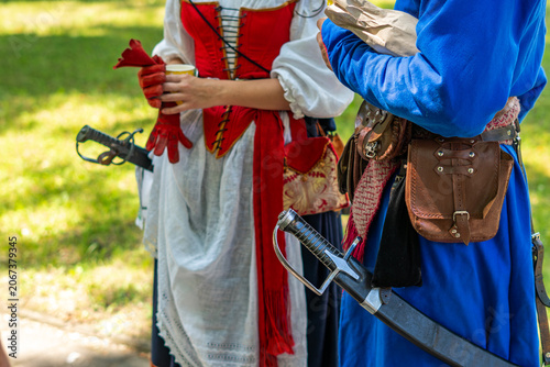 Photos Close-up of people in historical reenactment costumes, showing details of a woman's red corset with a white apron and a man's blue tunic with leather pouches and sabers in scabbards at the waist