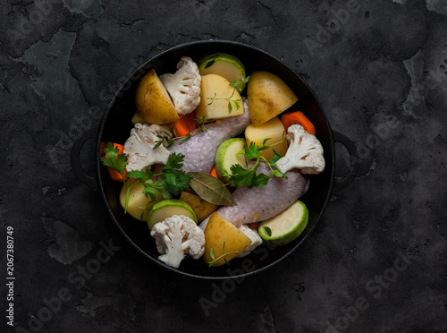 Slow cooking chicken drumsticks with vegetables in a cast-iron skillet on a dark background, top view. Raw food ingredients