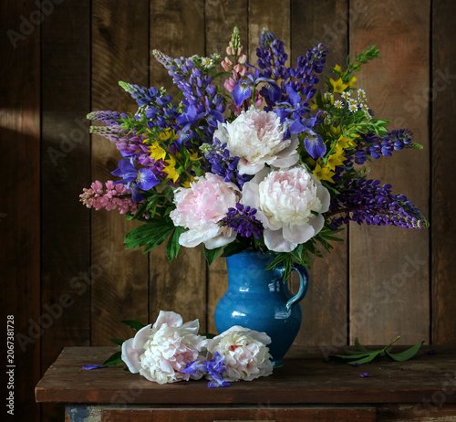 Rustic floral still life with peonies, irises, lupines and meadow flowers arranged in ceramic jug on dark wooden plank background.