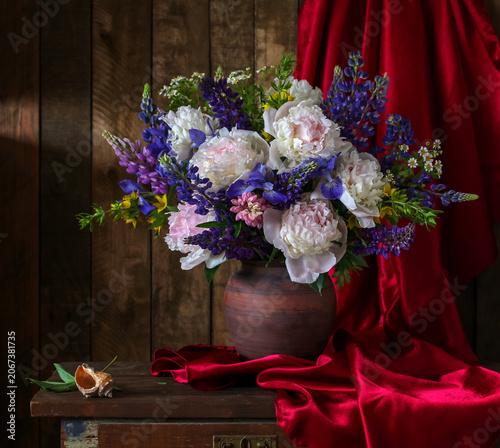 Rustic floral still life with peonies, irises, lupines and meadow flowers in clay jug with red satin drapery and shell, on dark wooden background.