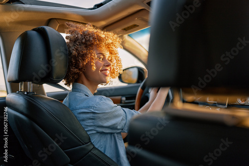 A smiling young woman with curly hair drives a car, wearing a denim shirt, sunlight on her face, capturing a candid moment of everyday mobility and independence.