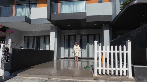 Woman performs exterior maintenance by hosing down dark patio tiles. Water spray from green garden hose washes townhouse yard, keeping modern architecture property clean and looking beautiful.