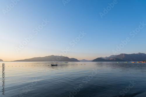 Scenic waterfront in Marmaris, Turkey during blue hour at sunrise or sunset. The coastal promenade overlooks calm sea framed by mountain silhouettes in the distance. Small boat floats gently on waves