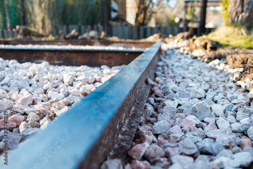 Close up, low angle view of steel edging on crushed pink white aggregate, set for a walkway foundation. Daylight scene shows sharp texture, blurred fence and trees.
