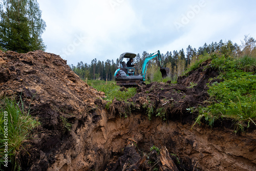 A compact teal excavator digs a wide pit in countryside, raw soil layers and roots dominate, operator works near a treeline under overcast daytime light.