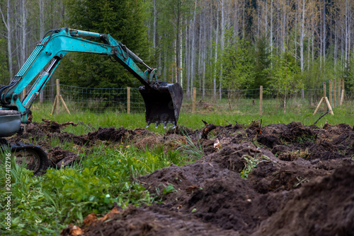 A teal mini excavator digs dark soil in a fenced woodland clearing, bucket mid scoop near fresh mounds and roots, spring saplings and soft daylight mark rural worksite.