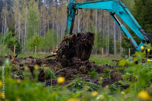 Foto A teal hydraulic excavator hoists wet soil and tangled roots in a rural field with wooden posts and mixed conifer and birch trees on an overcast day, low angle view