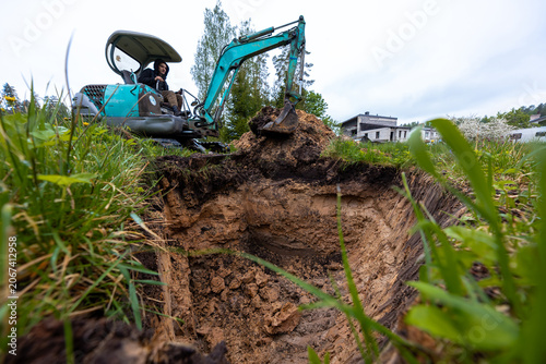 Konstfotografi Compact teal excavator with raised bucket works near a rectangular pit, layered soil and roots visible, operator in cab by a modern white building under overcast light