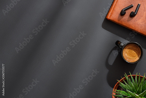 Fotografia Leather notebook with wireless earbuds, black coffee espresso cup and succulent plant on black background in dramatic soft lighting