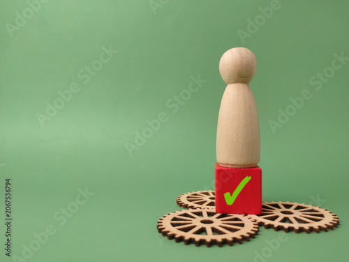 A wooden peg figure stands on a red checkmark block atop three interlocking wooden gear wheels on a green background