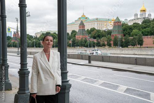 Photography Woman stands on Sofiyskaya Embankment in Moscow with Kremlin across river, framed by colonnade and overcast sky in contemporary urban setting