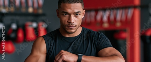 In a vibrant gym setting, an athlete stretches while glancing at a smartwatch, surrounded by boxing equipment and medals.