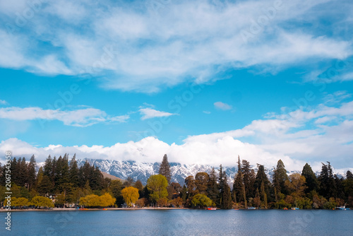 Queenstown, New Zealand: wide travel view of Lake Wakatipu waters with dense row of autumn foliage trees below snow-covered alpine mountain peaks.