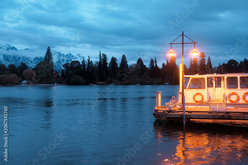 Queenstown, New Zealand: blue hour dusk travel view of illuminated yacht deck at wooden pier on Lake Wakatipu with warm lantern light reflection.
