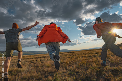 Low angle rear view of three young friends with backpacks running on green mountain meadow at sunset. Group of travelers celebrating success in remote scenic steppe wilderness.